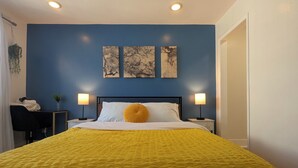 Une chambre avec un mur bleu, un lit recouvert d’une couvre-lit jaune, deux lampes de chevet et trois œuvres d’art accrochées au mur.