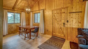 A wooden cabin interior with a dining table, two windows, a chandelier, and a door.