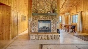 A log cabin interior with a stone fireplace, wooden walls, a dining area, and a large window.