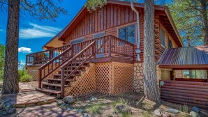 A log cabin with a wooden staircase leading to a balcony, surrounded by trees and a stone wall.