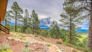A stone pathway leading to a wooden staircase in a mountainous area with pine trees and a view of distant mountains.