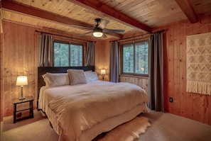 Quarto aconchegante com cama, mesa de cabeceira, abajur, ventoinha de teto e janelas amplas com cortinas.