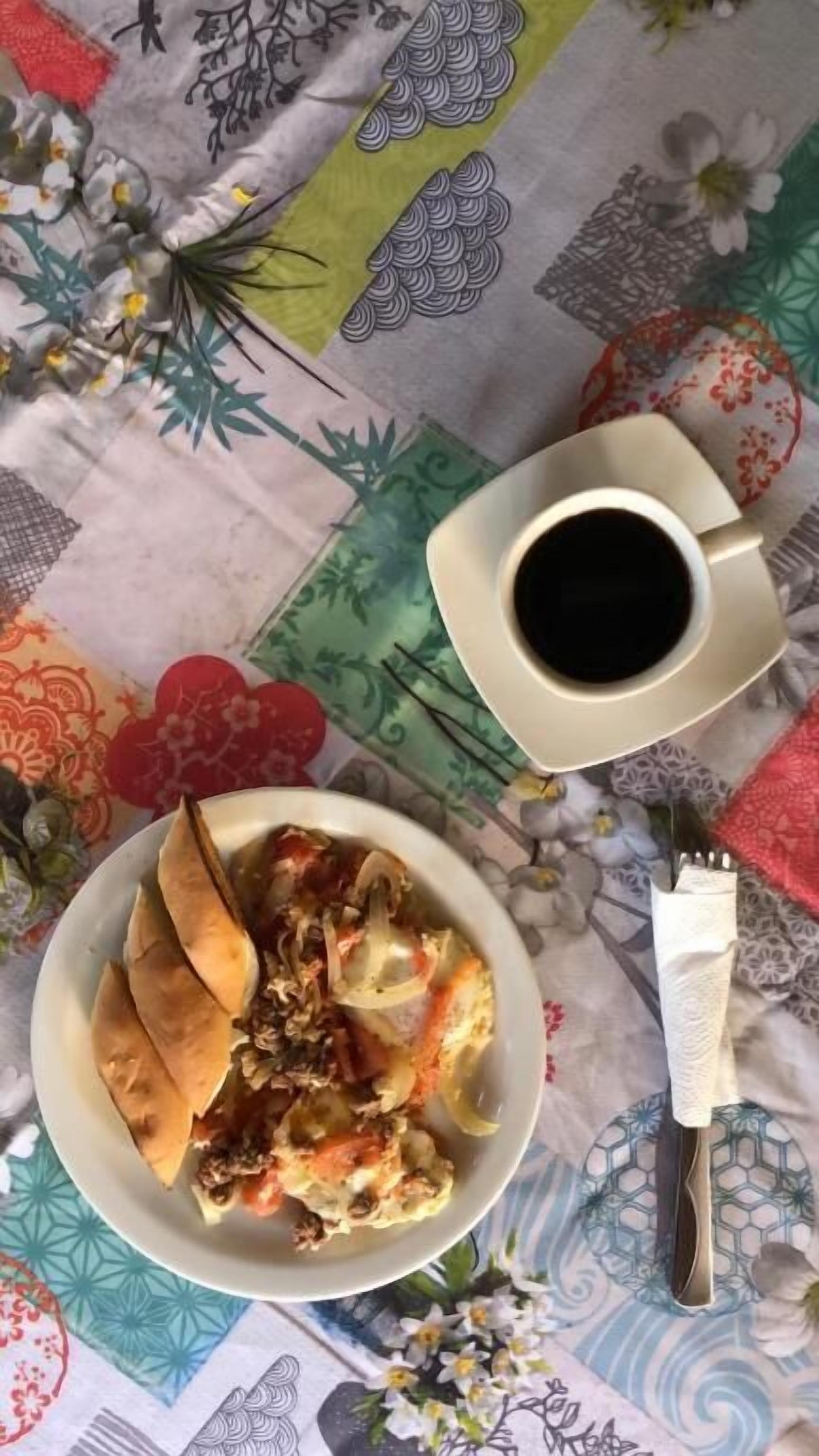 Una mesa con mantel floral, un plato de comida, una taza de café y una servilleta.