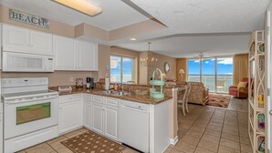 A modern kitchen with white cabinets, a microwave, stove, and dishwasher. There is a dining area with a chandelier and a view of the ocean. - Crescent Shores 1806 - Oceanfront condo rental  (North Myrtle Beach)