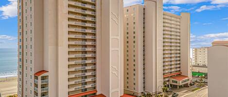 Un hotel de altura con varios balcones, vista a la playa y un estacionamiento con una carretera en primer plano.