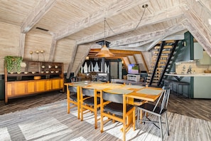 Un loft spacieux avec de hauts plafonds, un coin repas avec une table et des chaises en bois, une cuisine dotée d’un îlot central vert et un escalier menant à un niveau supérieur.