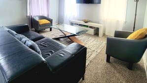 Ett vardagsrum med en svart lädersoffa, ett soffbord i glas, en vit TV-bänk, en golvlampa och en tavla på väggen.