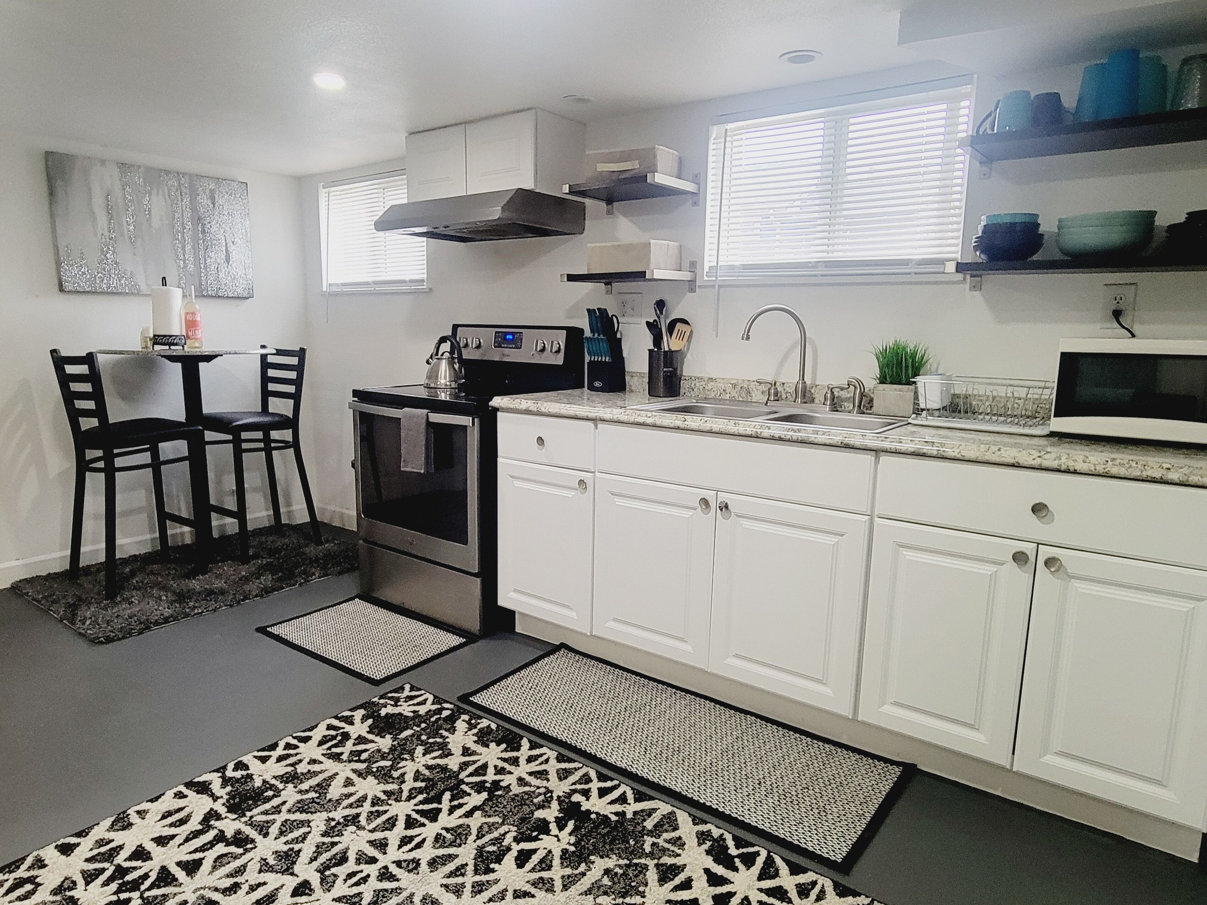 A modern kitchen with white cabinets, a stainless steel stove, a microwave, and a sink. There are two black chairs and a small table with a kettle.