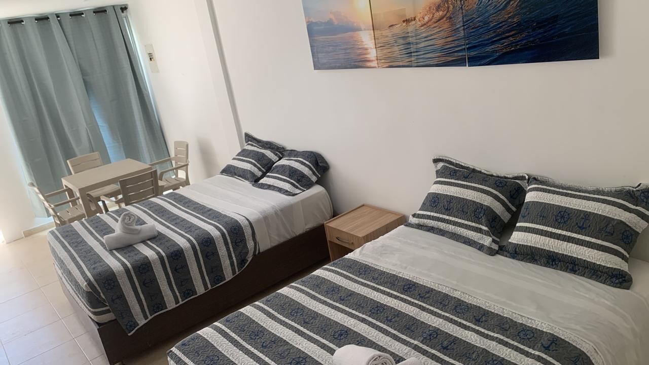 Un dormitorio con dos camas, una mesita de noche de madera, una pequeña mesa con sillas y un amplio ventanal con cortinas.