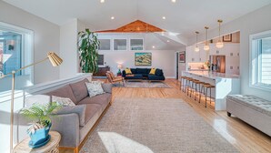 En rummelig opholdsstue med sofa, sofabord og en stor plante. Der er et køkkenområde med barstole og et spisebord.