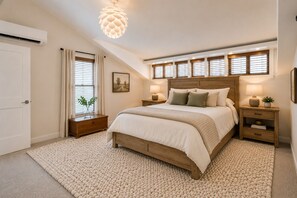 Une chambre à coucher comprenant un lit, des tables de chevet, une fenêtre avec des stores, un luminaire suspendu et une porte.