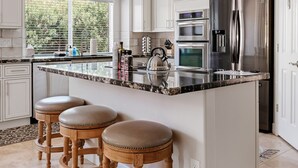 A modern kitchen with white cabinetry, a large island with stools, stainless steel appliances, and a window with a view of greenery.