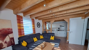 A living room with a dark blue sofa, wooden coffee table, and yellow cushions. A kitchen is visible in the background. - HM - Holiday home 3 Deluxe Krombachtalsperre Westerwald (Driedorf)