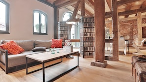 Ein geräumiges Wohnzimmer mit Holzboden, einem Sofa mit orangefarbenen Kissen, einem Couchtisch und einem Bücherregal voller Bücher.
