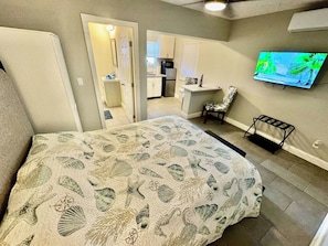 A bedroom with a bed, a TV, a chair, and a kitchenette in the background.