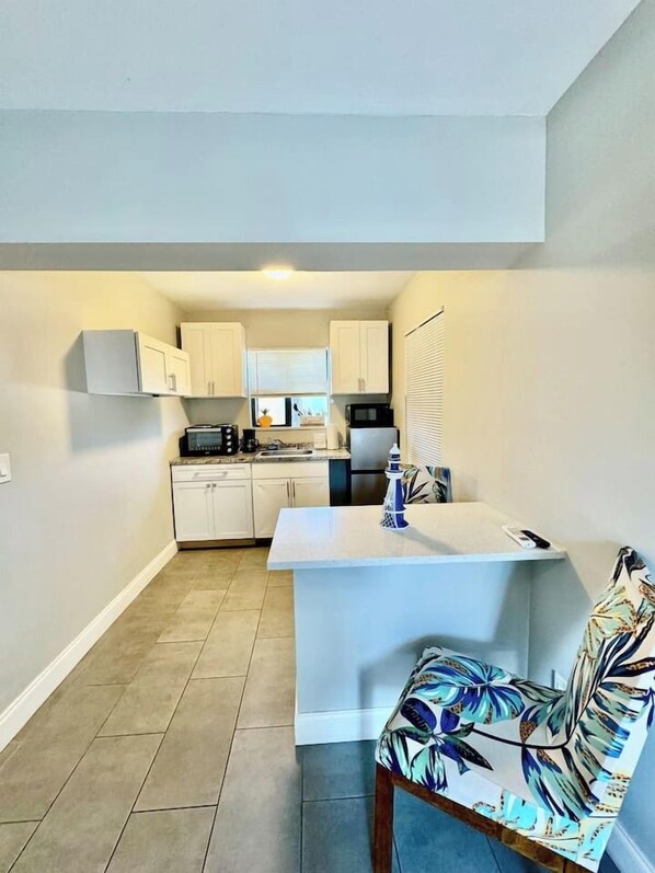 A compact kitchen with white cabinets, a microwave, and a small dining area with a table and a patterned chair.