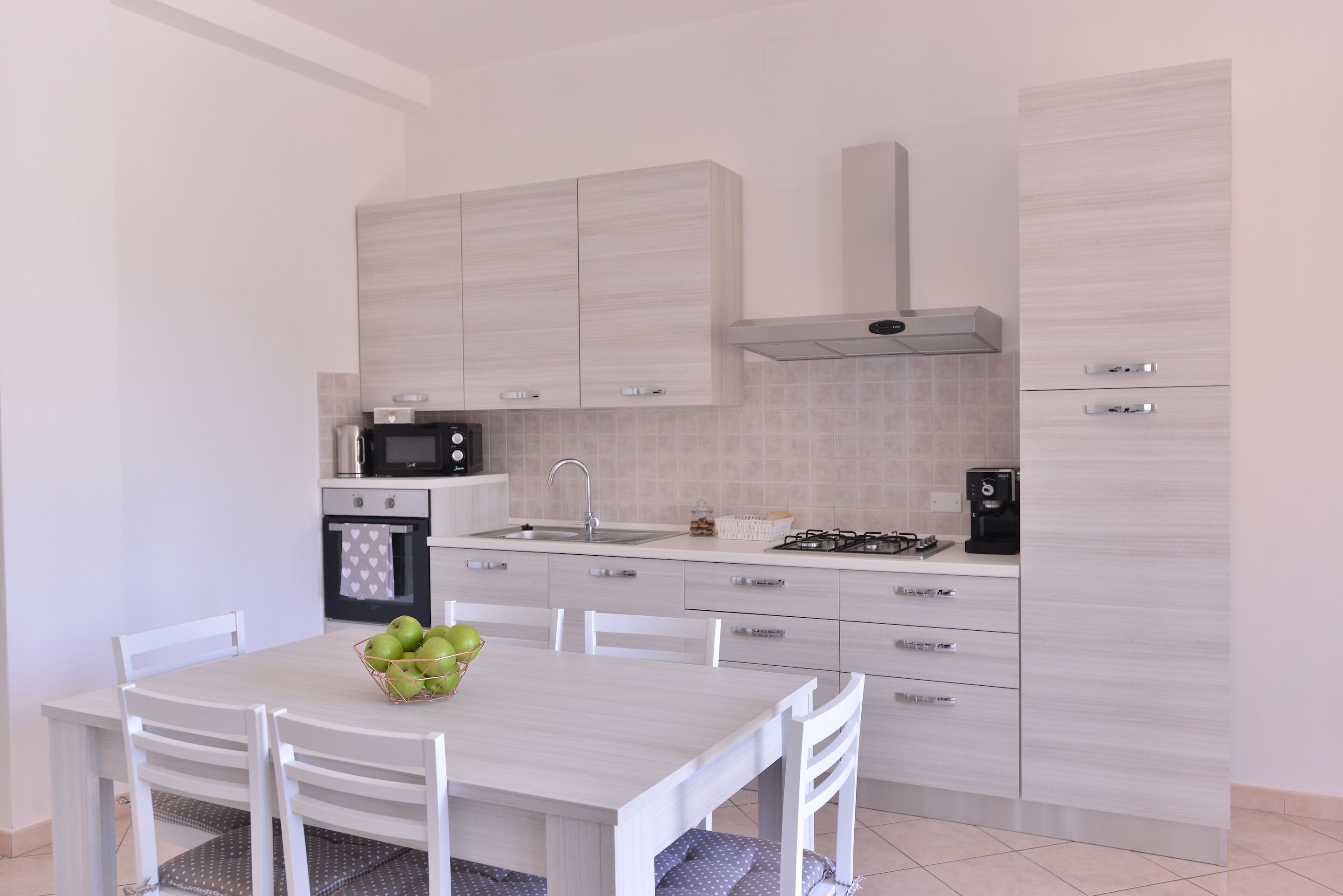 Una cucina moderna con mobili bianchi, un fornello a gas, un forno a microonde e un lavandino. C'è un tavolo da pranzo con sedie e una ciotola di mele verdi.
