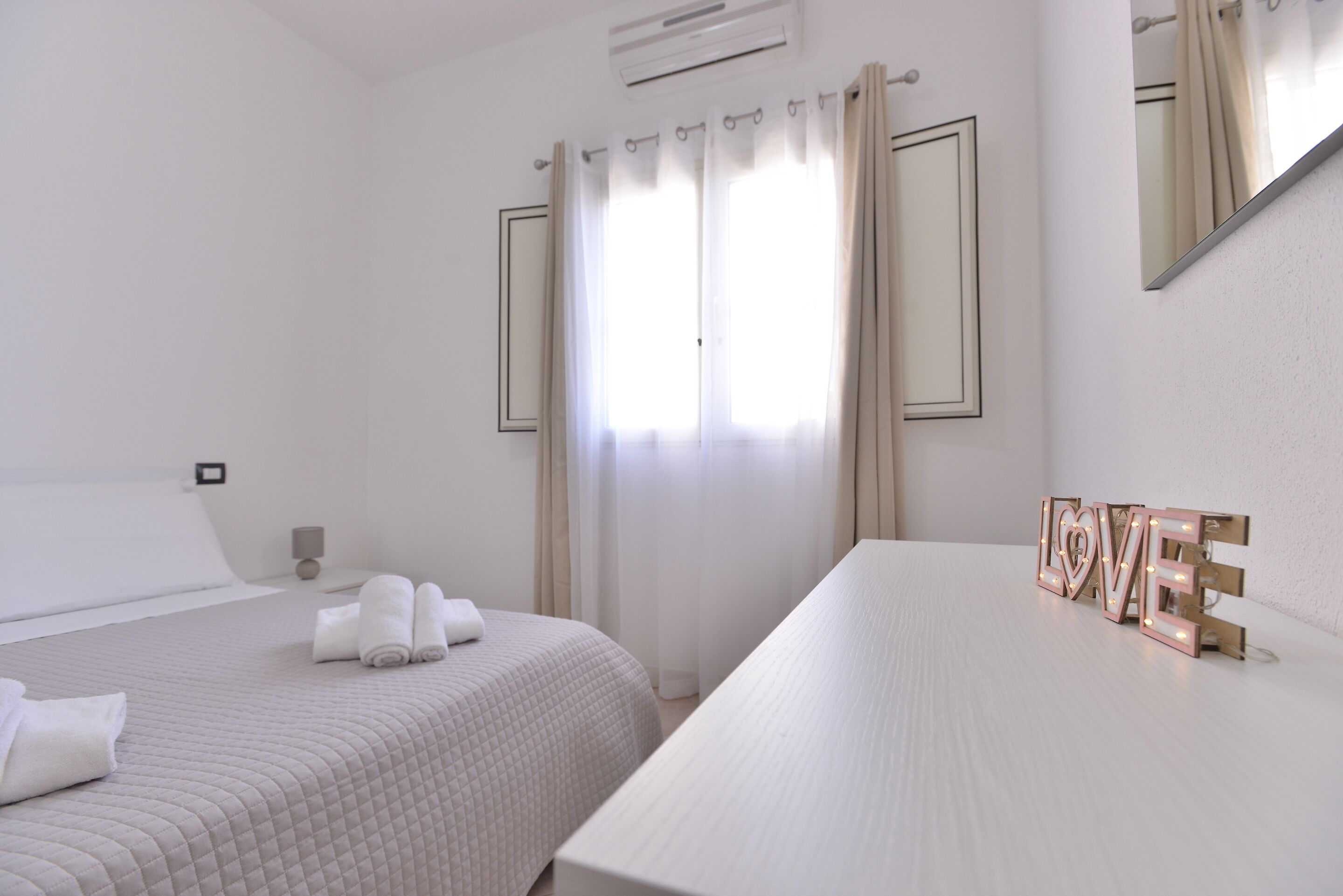 A hotel room with two beds, a bedside table, a mirror, and a large window with curtains.