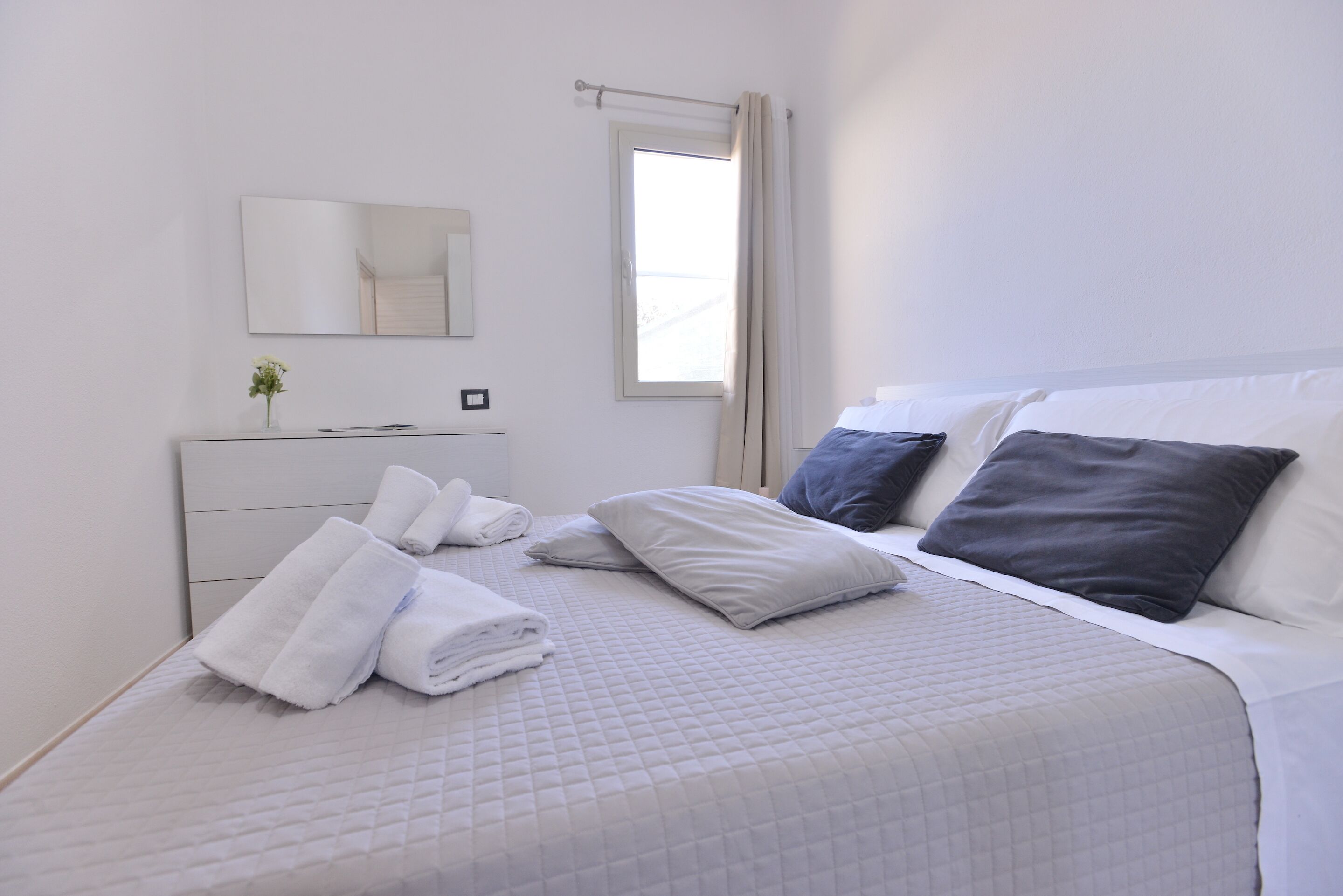 A neatly made bed with white and grey bedding, a bedside table with a vase of flowers, and a window with curtains.