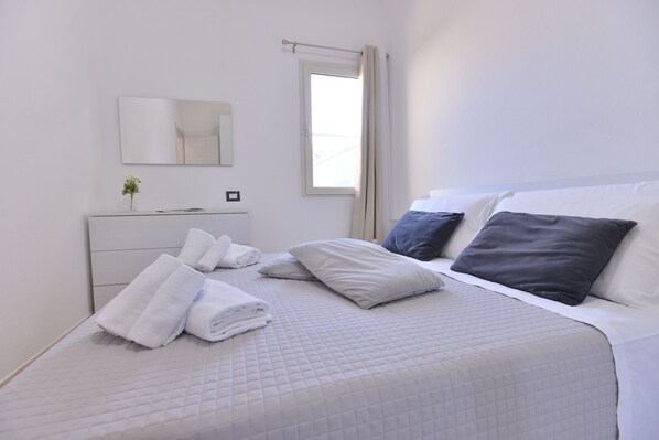 A neatly made bed with white and grey bedding, a bedside table with a vase of flowers, and a window with curtains.
