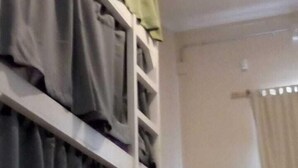 A room with a built-in shelving unit, a bed with a headboard, and a window with curtains.