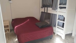 A small, single-bed room with a red bedspread, a wooden nightstand, and a white bunk bed with grey curtains.