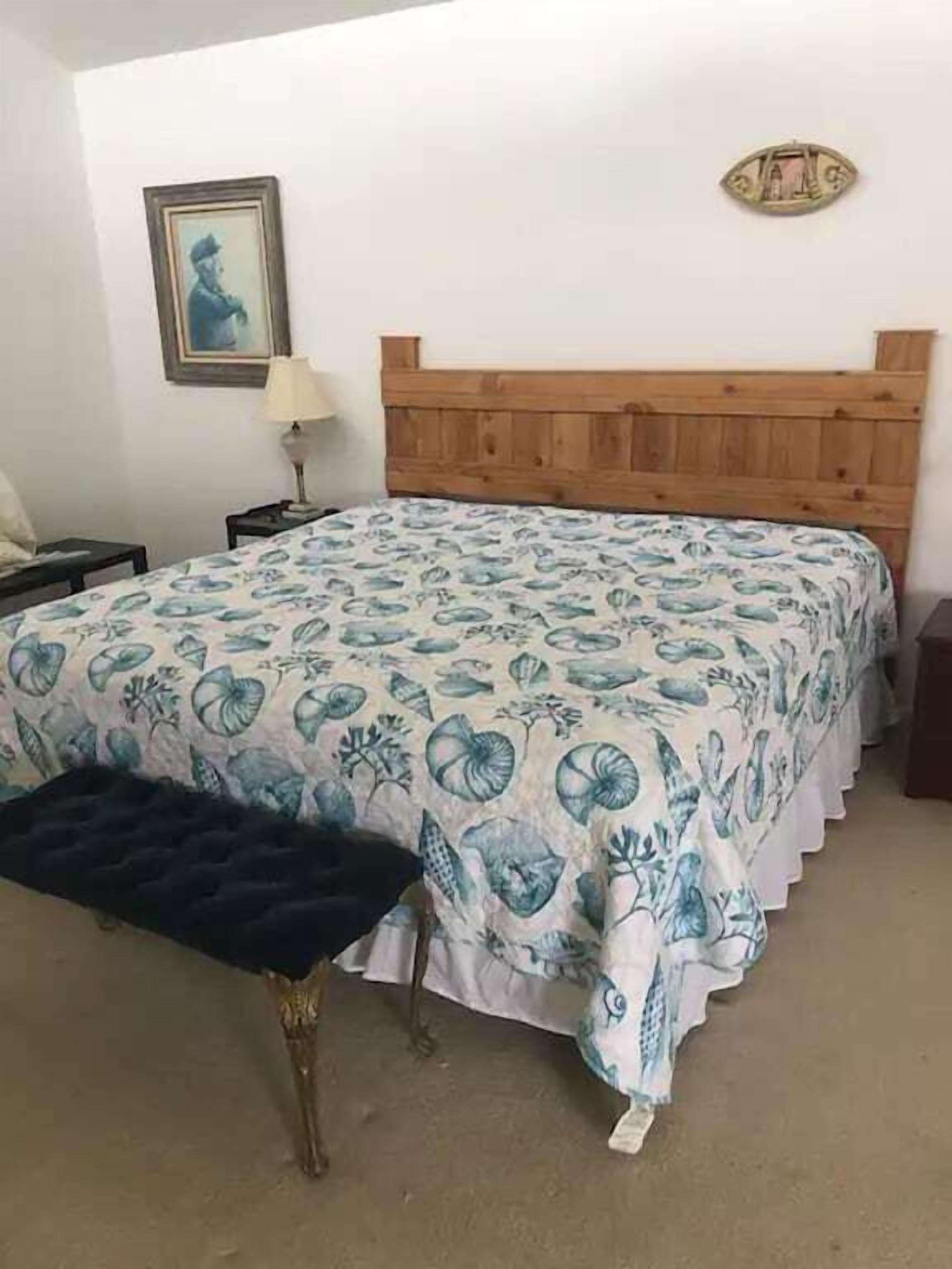 A bedroom with a wooden headboard, a floral bedspread, a bench, a nightstand, a lamp, and a framed picture on the wall.