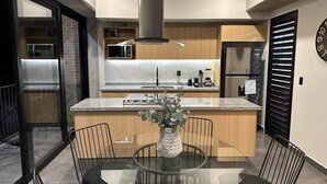 Una cocina moderna con una isla central, un área de comedor con mesa de vidrio y un ventanal grande.