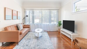 Un saló amb un sofà de cuir marró, una taula de cafè blanca, un televisor de pantalla plana sobre un suport blanc i un finestral gran amb cortines primes.