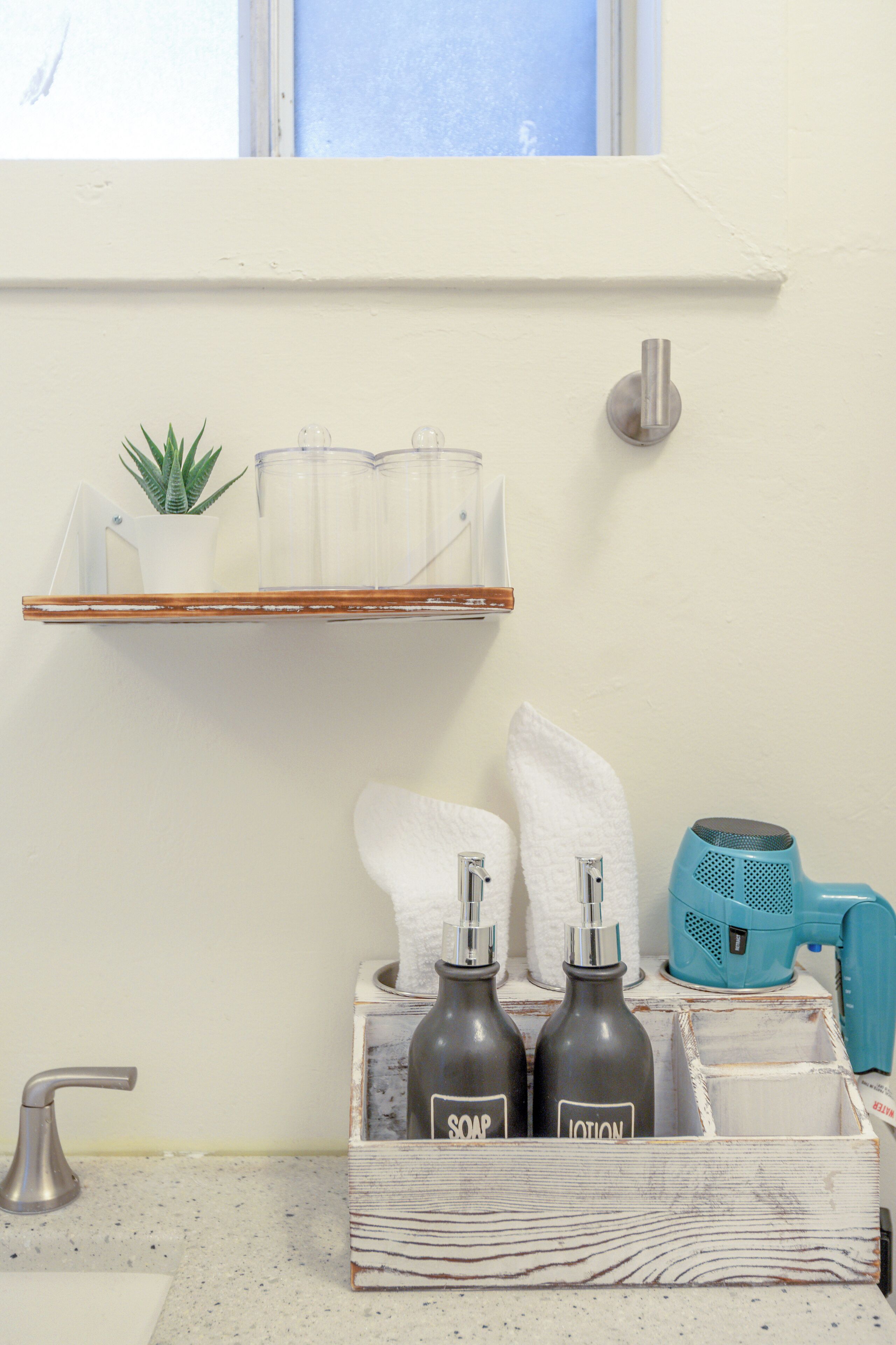 A bathroom with a shelf holding a plant, a glass container, and a towel. Below, there are bottles labeled 'SOAP' and 'LOTION' and a blue hairdryer.