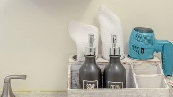 A bathroom with a shelf holding a plant, a glass container, and a towel. Below, there are bottles labeled 'SOAP' and 'LOTION' and a blue hairdryer.