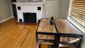 Una sala de estar con chimenea, un televisor montado encima, una mesa de comedor con sillas y una ventana con persianas.