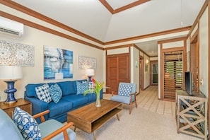 A living room with a blue sofa, wooden coffee table, and patterned cushions. There is a TV stand with a TV and a decorative vase on the coffee table.