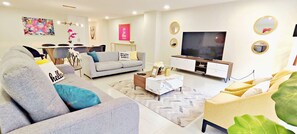 A modern living room with a grey sofa, a coffee table, and a TV. There is a dining area in the background with a table and chairs.