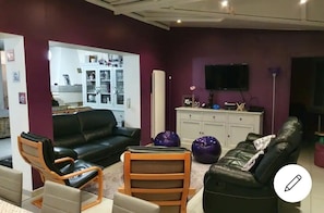 A living room with a black leather sofa, wooden chairs, a cabinet with decorative items, and a television on the wall.