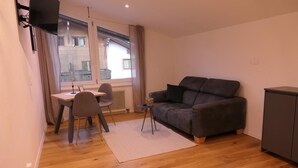 A compact living space with a wooden floor, a grey sofa, a small dining table with two chairs, and a radiator near the window. - Newly renovated, cozy apartment with a beautiful balcony - NEST'L Appartements (Mösern)