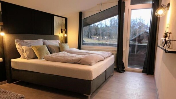 A modern bedroom with a large bed, a window with a view of trees, and a small table with a plant. - Newly renovated, cozy apartment with a beautiful balcony - NEST'L Appartements (Mösern)