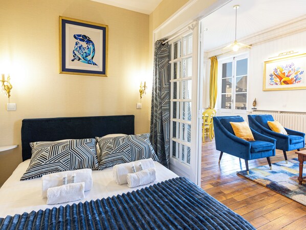 A hotel room with a bed, two chairs, a dining area, and artwork on the walls.