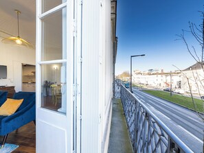 Un balcon donnant sur une rue, un coin repas avec un canapé bleu, et une cuisine visible à travers une porte ouverte.