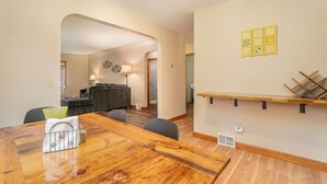 Un comedor con una mesa de madera, sillas, una pequeña cocina al fondo y un estante montado en la pared con objetos decorativos.