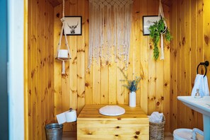 Una habitación de madera con un banco de madera, un tapiz macramé en la pared, un cuadro enmarcado, un jarrón blanco con plantas, un cubo de metal y un lavabo.