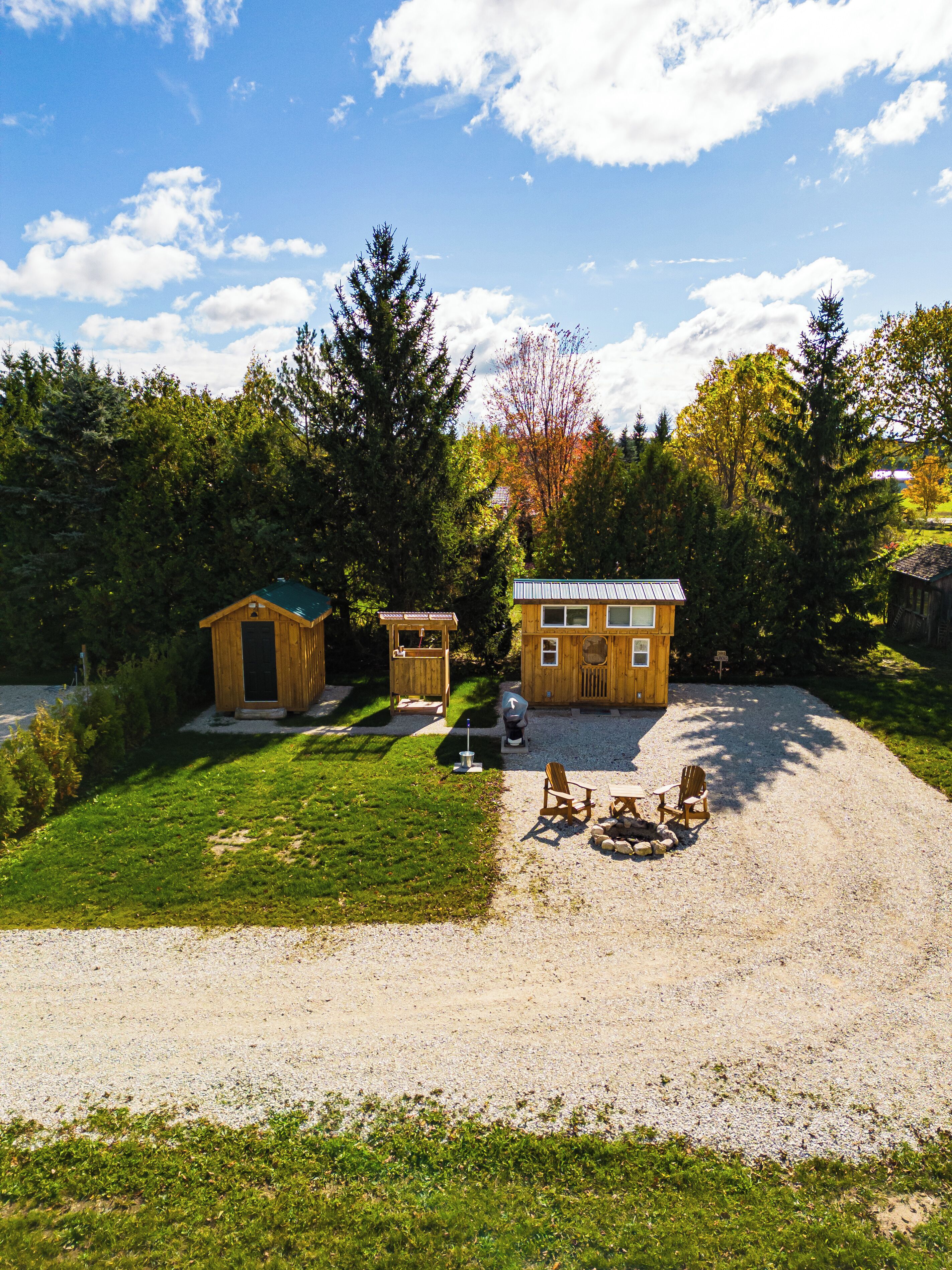 A wooden cabin with a green roof, a small shed, and a gravel driveway with two Adirondack chairs.