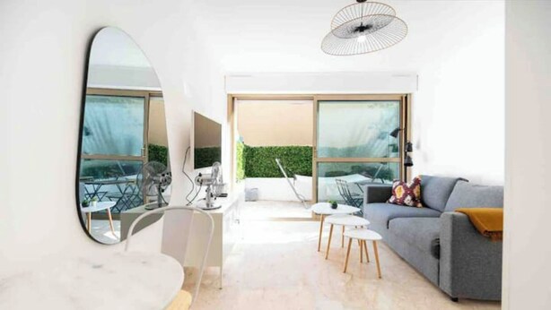 A modern living room with a grey sofa, a round table, and a large mirror. There is a balcony with a view of greenery. - Nice: Superb apartment in the Carré d'Or. (Nice)