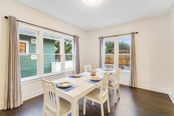 Uma sala de jantar com uma mesa branca posta para quatro pessoas, grandes janelas com cortinas e vista para uma casa verde e árvores.
