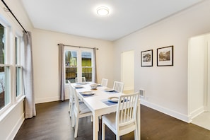 Uma sala de jantar com uma mesa branca posta para seis pessoas, cadeiras brancas e uma janela com cortinas.