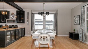 A modern kitchen with a central island, a dining area with a white table and chairs, and a large window offering a view of the city.