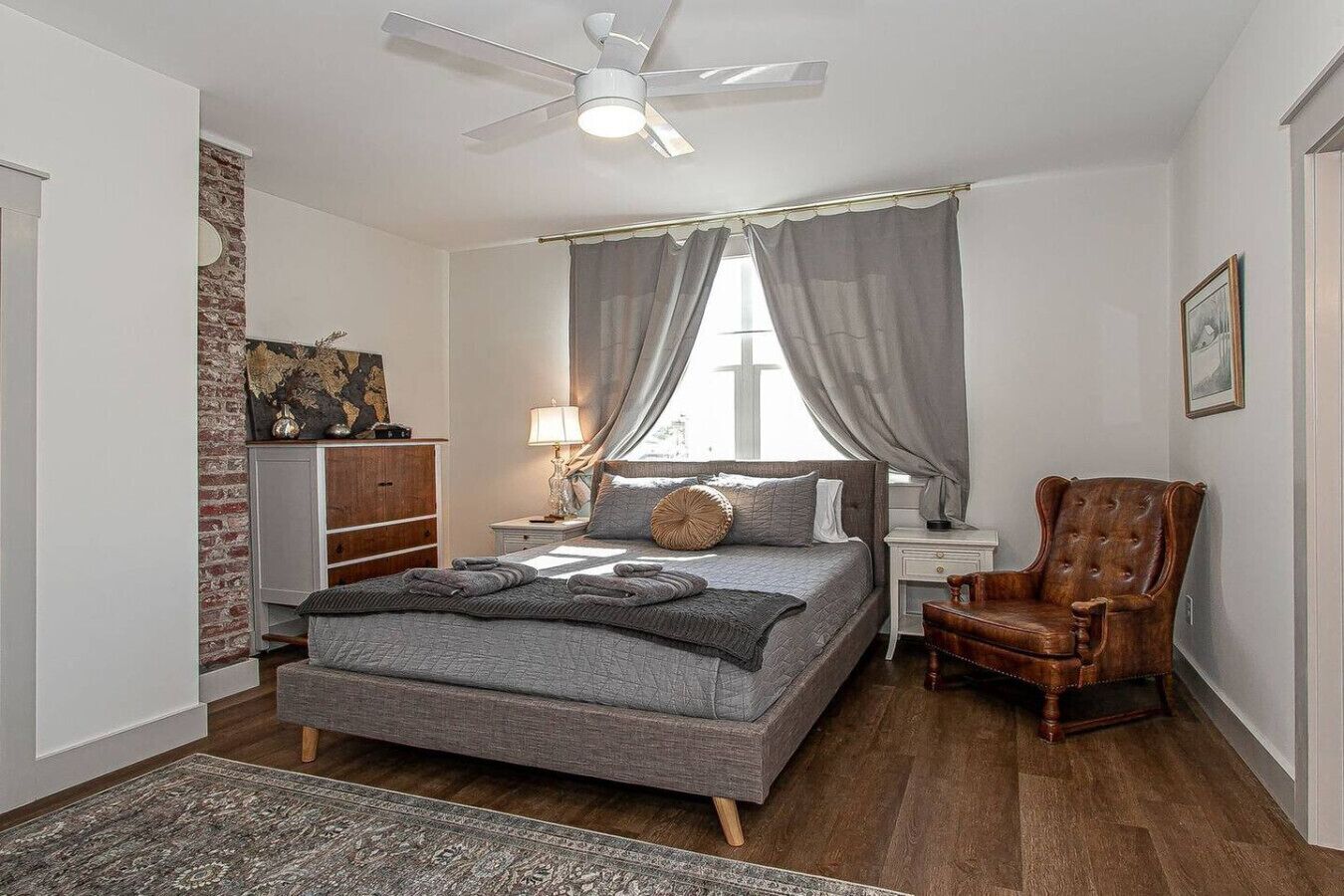 A bedroom with a bed, a chair, a nightstand, a lamp, a ceiling fan, and a window with curtains.
