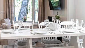 Un coin repas avec une table blanche dressée pour quatre personnes, un téléviseur fixé au mur et une fenêtre avec des rideaux.