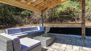 Un espace extérieur aménagé, abrité et doté d'un canapé en osier et d'une table en verre, entouré d'un dallage en pierre et d'une pergola en bois.