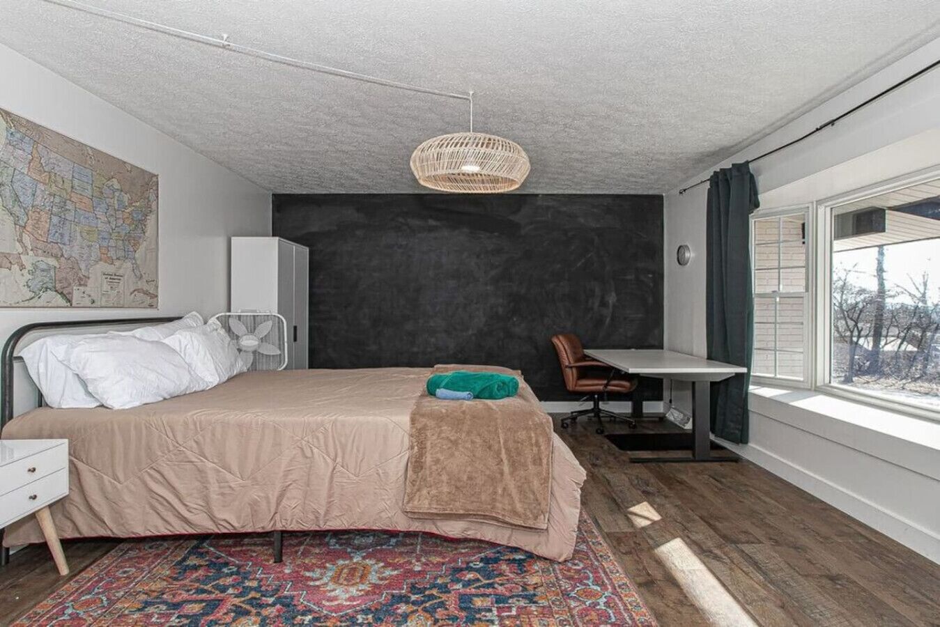 A bedroom with a bed, a desk, a chair, a map on the wall, and a window with a view of trees.